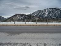 snow covering the ground with mountains in the background on a cloudy day, near an empty road and fence