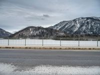 snow covering the ground with mountains in the background on a cloudy day, near an empty road and fence