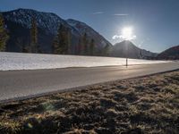 the road runs between two snowy mountains with low snow on them under the sun flare