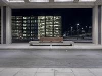 the empty city street has a bench by it's curb by a building at night