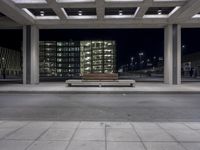 the empty city street has a bench by it's curb by a building at night