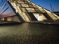 an illuminated building at night on a cobblestone floor, with a stair case and glass front