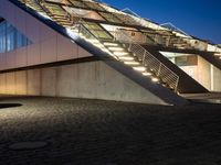 an illuminated building at night on a cobblestone floor, with a stair case and glass front