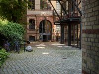 bicycles parked on a brick walkway between buildings and trees that are closed off in the shade