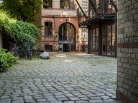 bicycles parked on a brick walkway between buildings and trees that are closed off in the shade