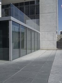 the glass enclosed building has stairs and a railing on one side of it and a concrete building behind it