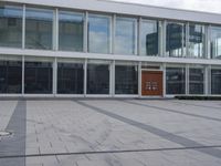 a brick walkway in front of a large building, with glass windows and doors along the front