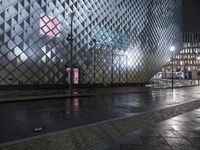 people walking down a sidewalk near a train station at night in the rain, with lights shining on them