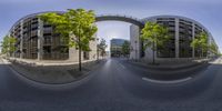 a street with trees, a building and an arch between them in a fisheye lens