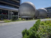 three large metallic spheres in front of a building in a city with tall buildings and bushes on the side