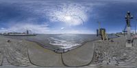 a panoramic view shows an old building and the ocean while people walk along a cobble stone street next to it