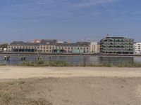 two people walk down a dirt path beside the water, in front of a row of buildings