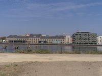 two people walk down a dirt path beside the water, in front of a row of buildings