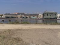 two people walk down a dirt path beside the water, in front of a row of buildings