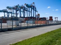 container terminal with containers and green grass on the side of a road near a fence and some tall cranes