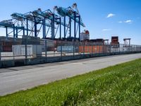 container terminal with containers and green grass on the side of a road near a fence and some tall cranes