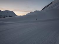 a person skiing down a snowy hill at dusk with a sky background and mountain range in the background
