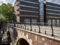 a brick walkway crosses an arched bridge over the water in a city with large buildings behind