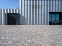 a brick walkway leading to a metal door in front of a building with a wall covered in vertical stripes