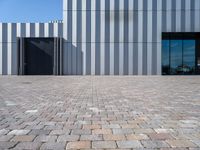 a brick walkway leading to a metal door in front of a building with a wall covered in vertical stripes