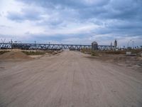 a dirt road that has a bridge in the background with a person on it on the other side