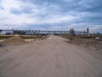 a dirt road that has a bridge in the background with a person on it on the other side