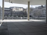 an empty concrete patio with white columns overlooking buildings and river in the middle of it