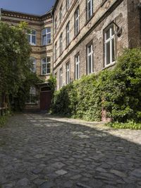 a cobble stone walkway next to buildings and bushes and trees and bushes next to an old brick building