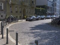 several parked cars on a cobblestone street near tall buildings with their shadows on the pavement