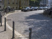 several parked cars on a cobblestone street near tall buildings with their shadows on the pavement