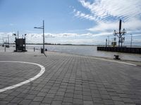 a parking area at the edge of a wide river near some poles and lights and a clock