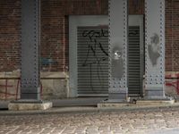graffiti on a metal door near an old building and a sidewalk with brick flooring