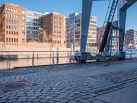 a person that is walking on brick pavement near water and buildings with a bridge in the background