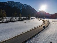 a highway runs past snowy mountains in a valley, with sun peeking over top of the mountain