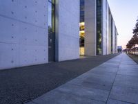 a walkway lined with grey cement next to an office building at dusk with light streaming in the windows