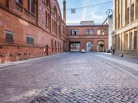 there is an alley with some brick buildings on both sides and a clock hanging over one of the cobblestones