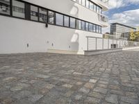 a skateboarder performs a trick on a block paved sidewalk outside a building,