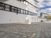 a skateboarder performs a trick on a block paved sidewalk outside a building,