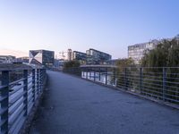 a walkway on a river bank between two buildings and a park area at sunset in the background