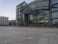 the outside of an architectural building with a few people inside it at dusk, on a plaza in front of a train station