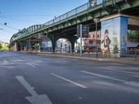 a view of an empty street, under an overhead bridge in a city, from the street with pedestrian and sidewalk markings