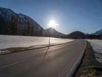 the sun shines brightly over a roadway in the mountains area near a snow covered field