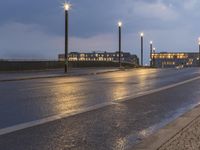 a wet street and some light poles on the side of the road at night with building lights