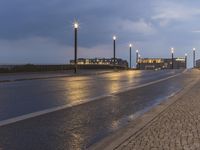 a wet street and some light poles on the side of the road at night with building lights