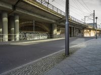 an overpass above a street with several graffiti on it's wall and a sidewalk with cobblestones