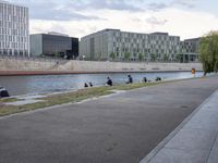 people sit on the shore next to the water in a city park next to the river and buildings