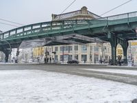 a street with a snowy road underneath the bridge and a snow - covered ground with a row of buildings in the background