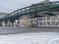 a street with a snowy road underneath the bridge and a snow - covered ground with a row of buildings in the background