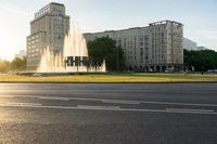 a water fountain spewing out from a green grass field next to some tall buildings