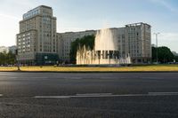 a water fountain spewing out from a green grass field next to some tall buildings